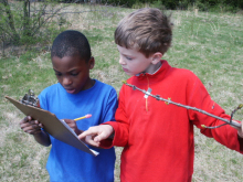 Two children looking at a clipboard and holding a stick.