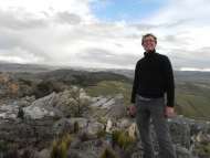 Bullard Fellow Robin Sears stands on a mountaintop in southern Peru, with rocks in the foreground and grass and forest-covered mountains in the distance