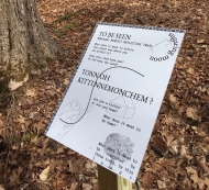 in the forest in early spring stands a white trail sign with black text that asks several questions including "are you a visitor, or are you home?"