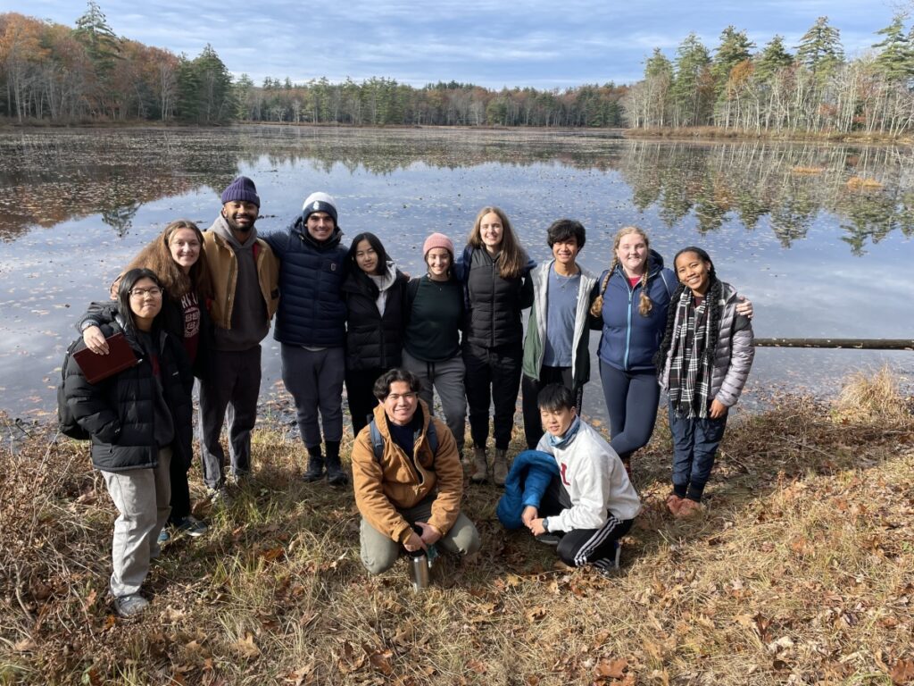 Eleven students and their proctor stand together on the shore of a pond