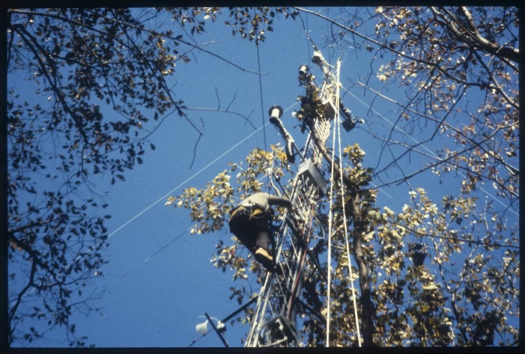 A man climbs a narrow steel tower that has scientific monitoring equipment mounted on it