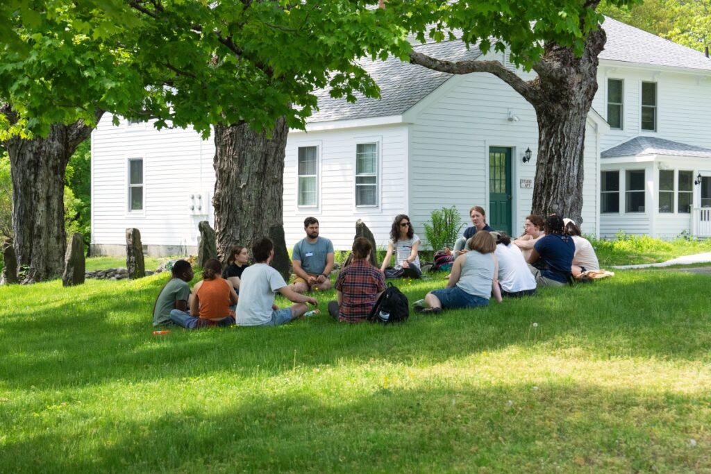 A group of students and their mentors sit on the lawn. A white house is in the background.