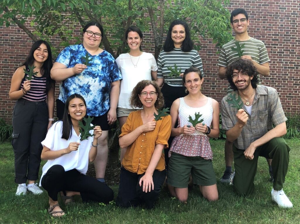 A group of 9 people holding oak leaves pose in front of a brick building