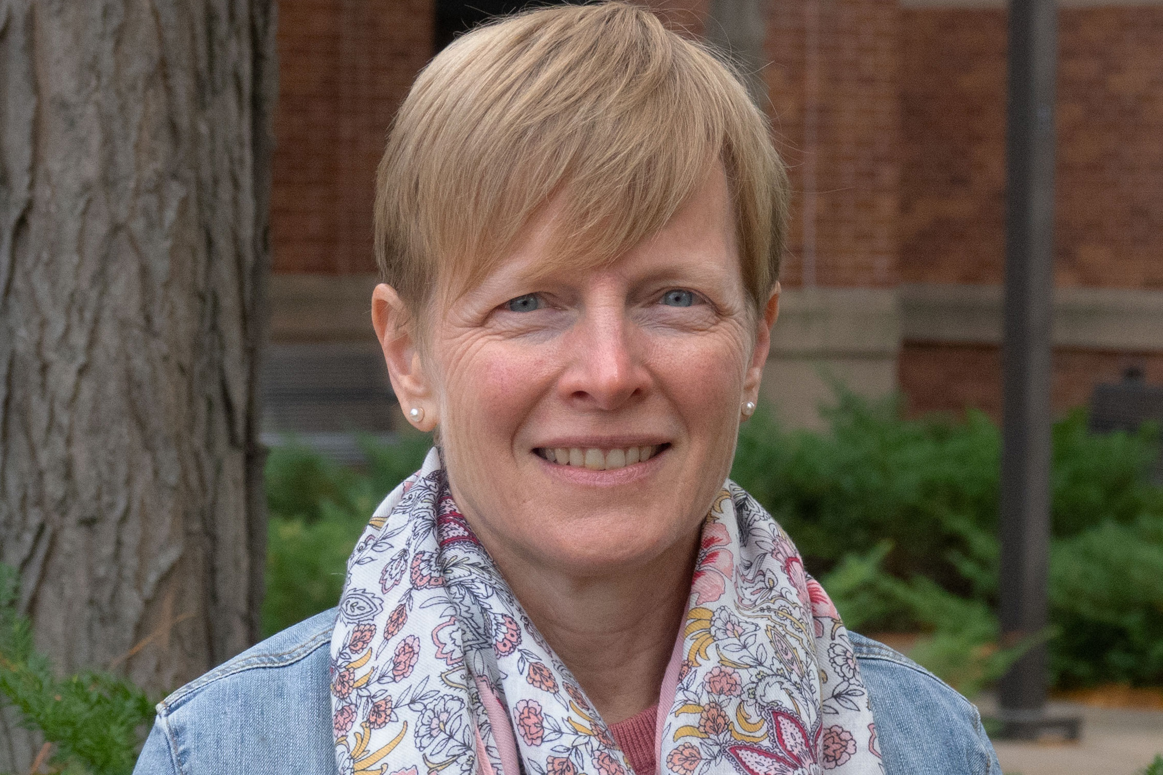 Close-up shows Sarah Hobbie, a smiling woman with short blonde hair, and a floral scarf, who is standing outside.