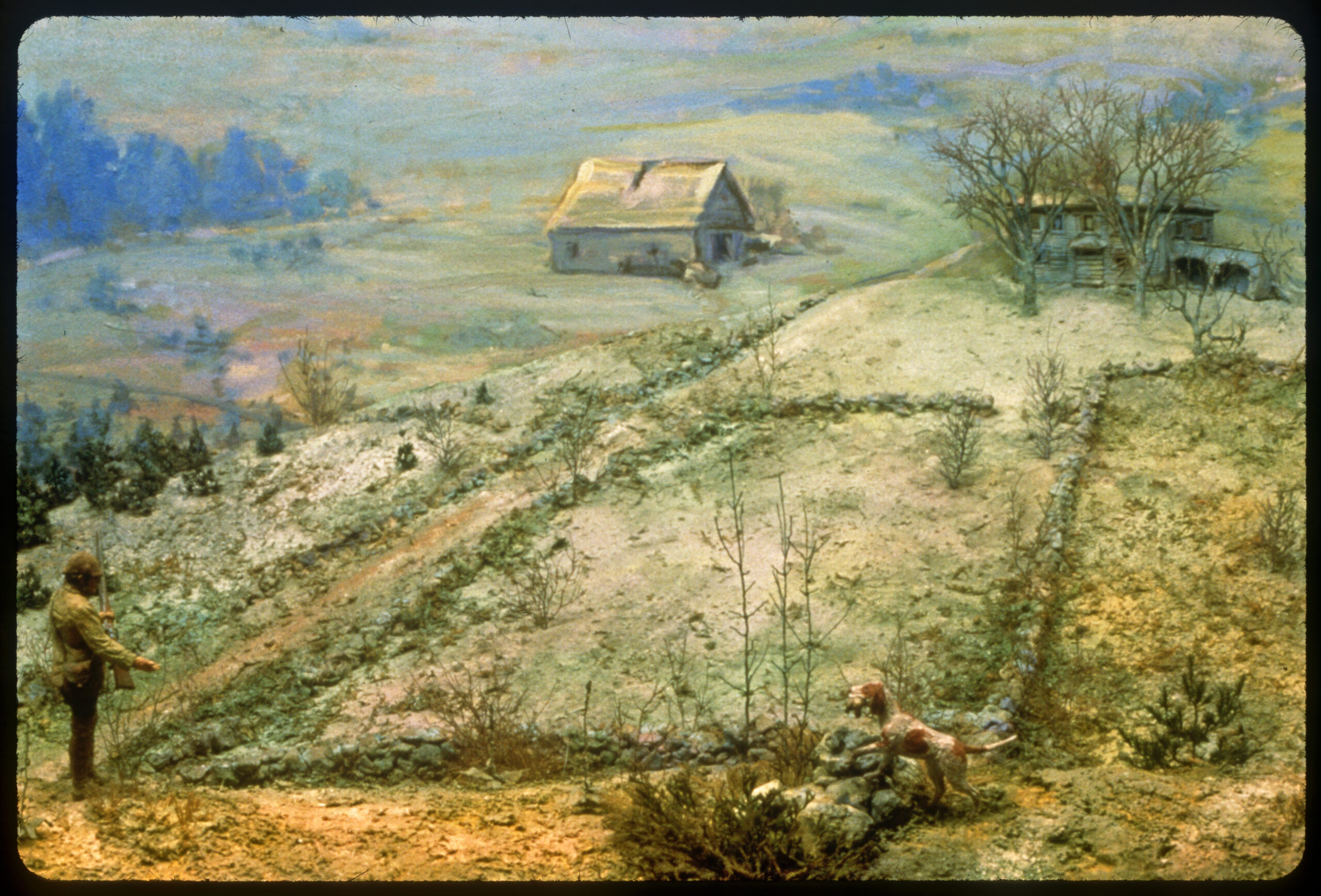 Diorama scene with an abandoned farmhouse and barn in the background. Small trees are beginning to grow in old fields. A hunter and his dog are in the foreground.