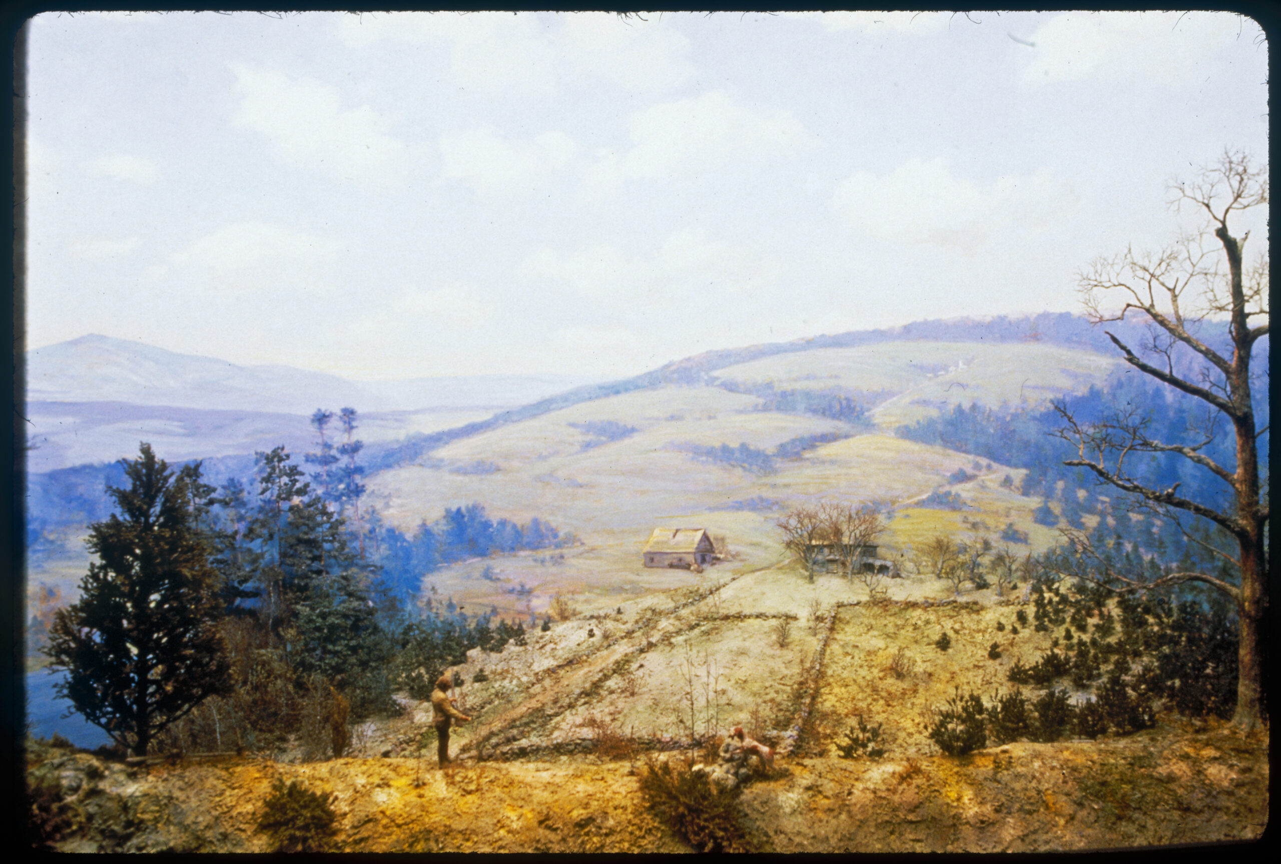Diorama scene with an abandoned farmhouse and barn in the background. Small trees are beginning to grow in old fields. A hunter and his dog are in the foreground.