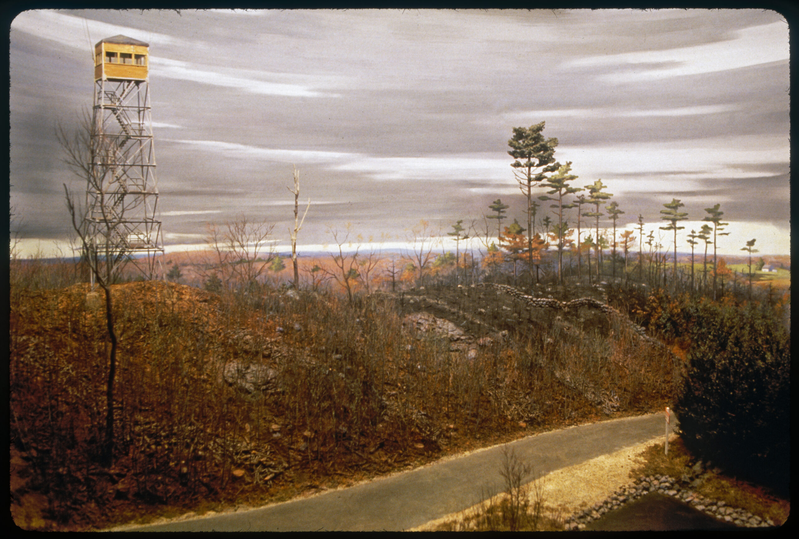 Diorama scene of a fire observation tower on a hill. A road runs through the scene and on the side of the road is a water hole.