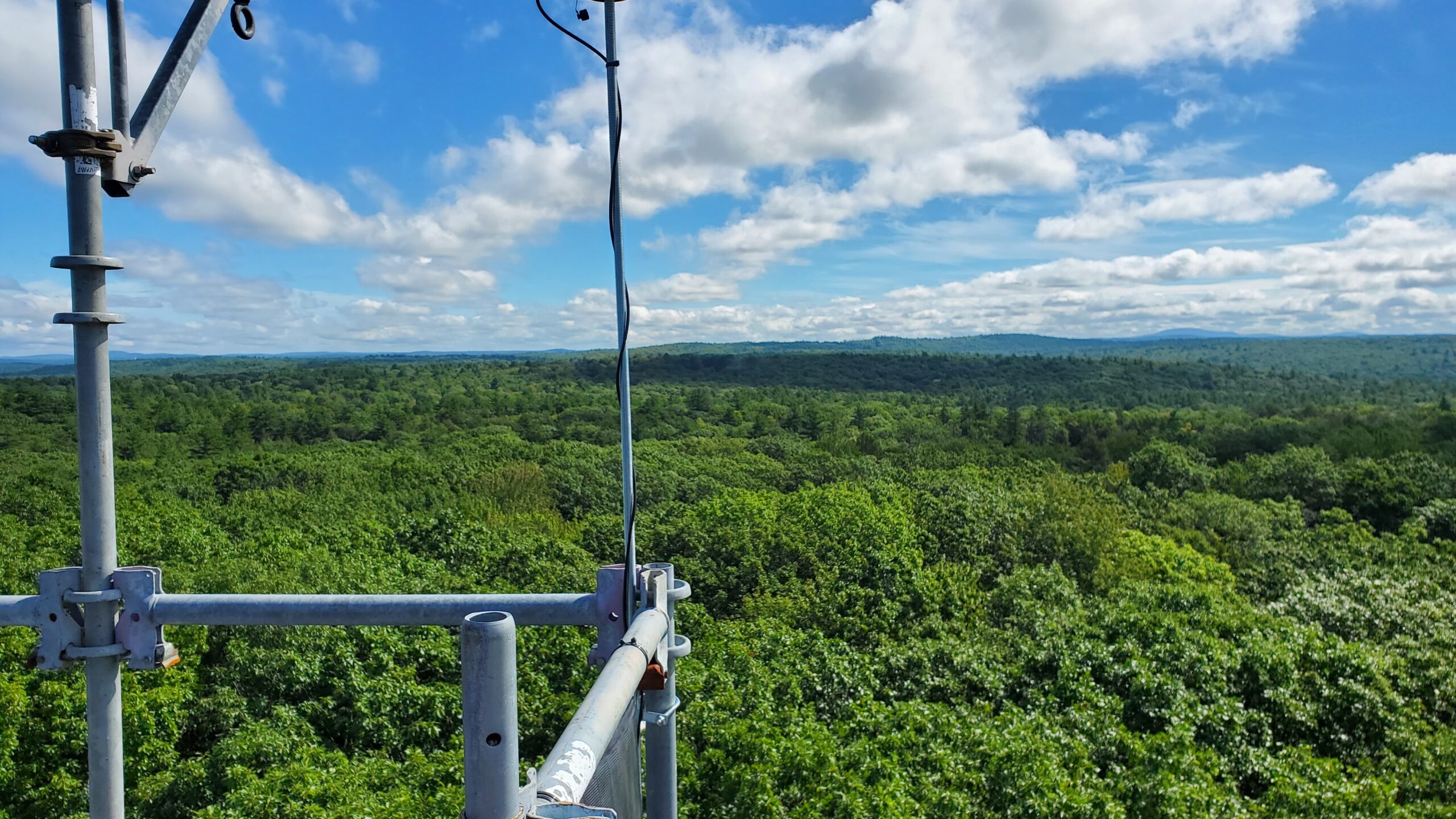 View of the sky and clouds from above the hardwood canopy with the tower railing and scientific equipment in the foreground