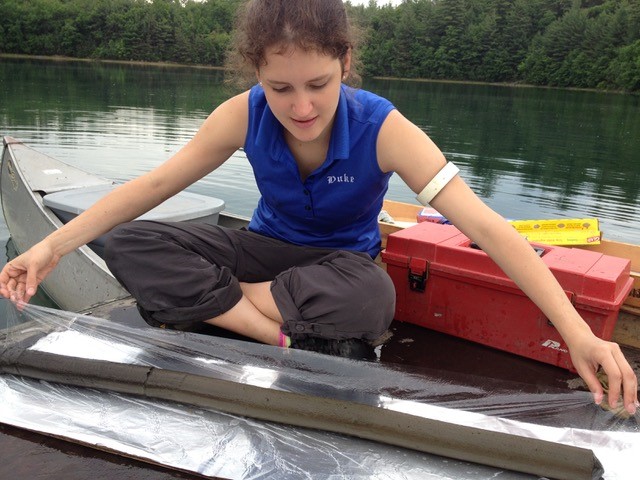 A student sitting cross-legged in a canoe wraps a cylindrical core of pond sediment in plastic wrap