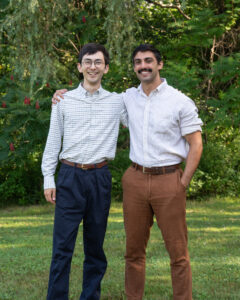 Ben and Jacob, two young men, stand smiling in button-down shirts in front of the green trees of a forest.