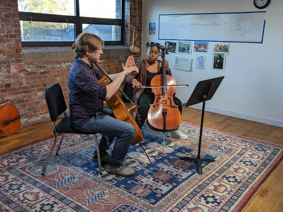 A young person with a cello engaged in a discussion with a teacher who also has a cello, indoors in a well lit room